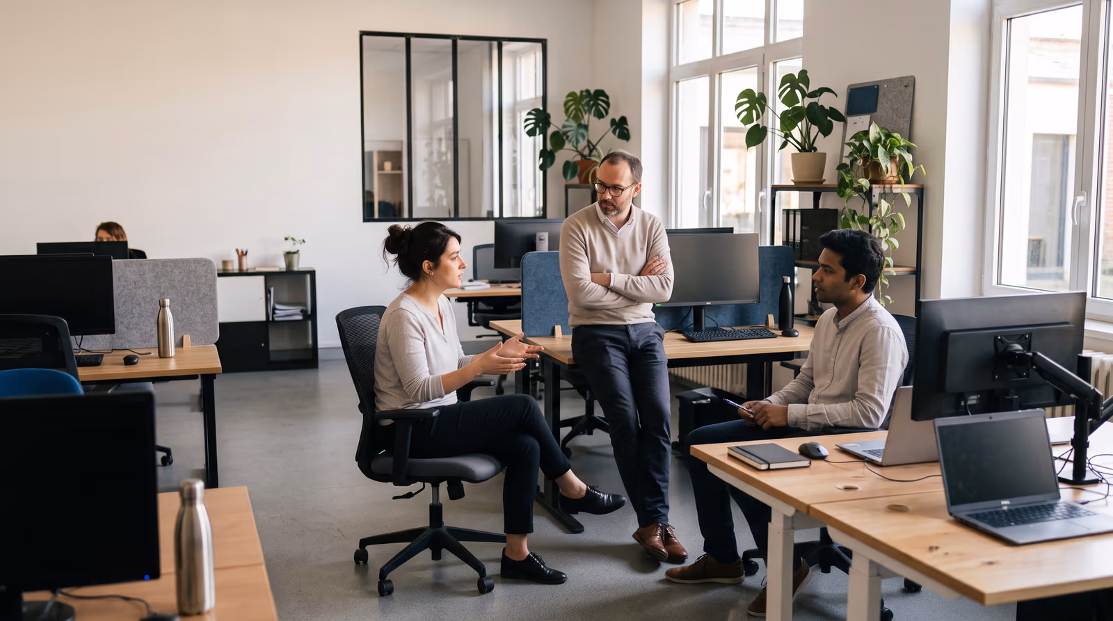 Three colleagues talking through a case in an open-plan office, one seated explaining, one leaning against a desk, one taking notes